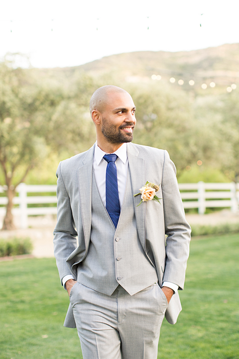 Los angeles outdoor wedding at brookview ranch groom heather grey notch lapel suit with a matching vest and white dress shirt and long blue tie and white floral boutonniere standing with hands in pockets