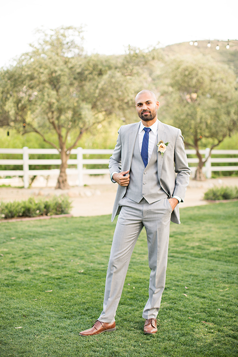 Los angeles outdoor wedding at brookview ranch groom heather grey notch lapel suit with a matching vest and white dress shirt and long blue tie and white floral boutonniere standing