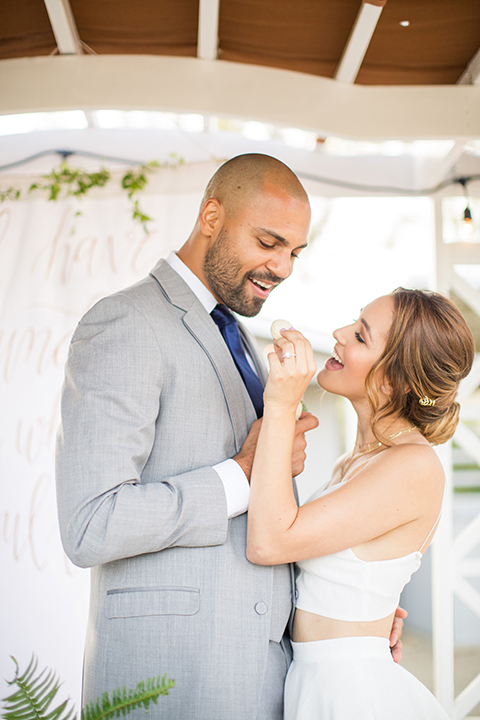 Los angeles outdoor wedding at brookview ranch bride two piece wedding dress with tulle skirt and crop top bodice with thin straps and a sweetheart neckline and groom heather grey notch lapel suit with a matching vest and white dress shirt with a long navy blue tie and white floral boutonniere hugging and eating cake by table