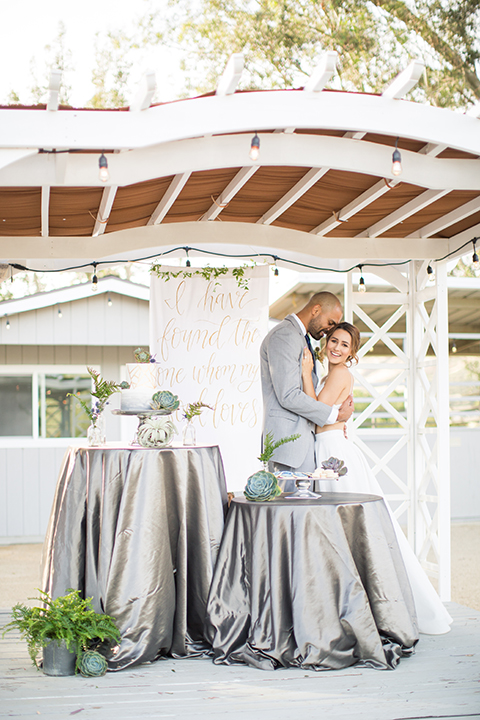 Los angeles outdoor wedding at brookview ranch bride two piece wedding dress with tulle skirt and crop top bodice with thin straps and a sweetheart neckline and groom heather grey notch lapel suit with a matching vest and white dress shirt with a long navy blue tie and white floral boutonniere hugging and standing by table