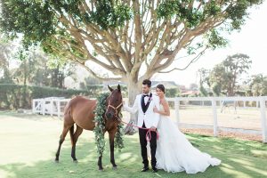 Santa barbara outdoor wedding at the cottages at polo run bride strapless ball gown with a sweetheart neckline and a ruffled skirt with lace detail on bodice with groom white shawl lapel tuxedo with black pants and a black vest with a black bow tie and bright pink floral boutonniere standing with horse