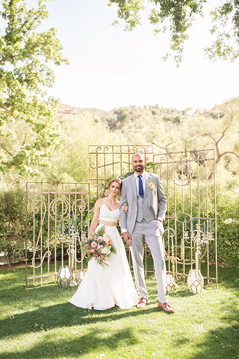 Los angeles outdoor wedding at brookview ranch bride two piece wedding dress with tulle skirt and crop top bodice with thin straps and a sweetheart neckline and groom heather grey notch lapel suit with a matching vest and white dress shirt with a long navy blue tie and white floral boutonniere hugging and holding hands during ceremony with backdrop and flower decor