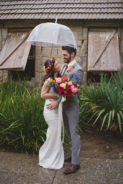 Temecula outdoor wedding at temecula creek inn bride form fitting simple strapless gown with a straight neckline and teal rain boots with groom grey notch lapel suit with a white dress shirt and long brown tie with an orange floral boutonniere standing holding clear umbrella and bride holding colorful floral bridal bouquet