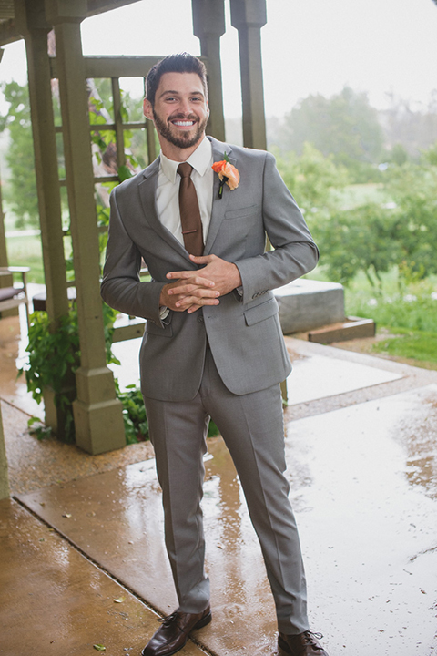 Temecula outdoor rainy wedding at temecula creek inn groom heather grey notch lapel suit with a white dress shirt and a long brown tie with a colorful floral boutonniere standing holding hands